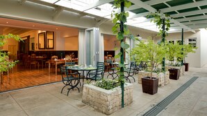 An indoor dining area with tables and chairs, a stone planter with a tree, and a view of the adjacent room. - Protea Hotel by Marriott Dorpshuis & Spa Stellenbosch (Stellenbosch)