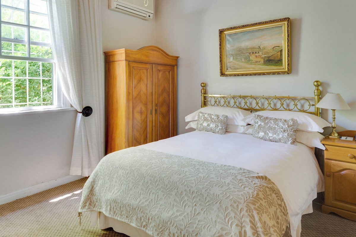 A bedroom with a bed, a wooden wardrobe, a nightstand with a lamp, and a painting on the wall.