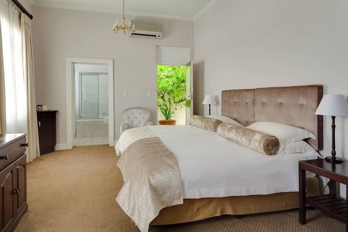 A bedroom with a large bed, a bedside table with a lamp, a chair, a dresser, and a door leading to a balcony with a view of greenery.