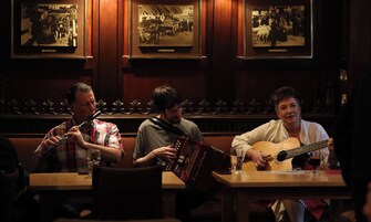 Tres músicos sentados a una mesa en una habitación tenuemente iluminada, con paneles de madera y fotografías históricas enmarcadas en la pared.