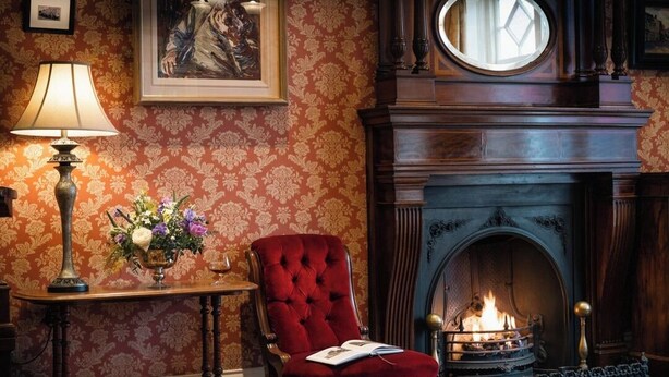 A room with a fireplace, a red armchair, a table with a lamp, and a vase of flowers. - Arbutus Hotel Killarney (Killarney)