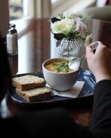 A bowl of soup with a spoon, two slices of bread on a tray, and a decorative vase with flowers in the background.