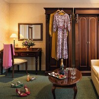 Una habitación con un escritorio de madera, una silla, un vestido colgado, un armario y una mesa con una botella y vasos.