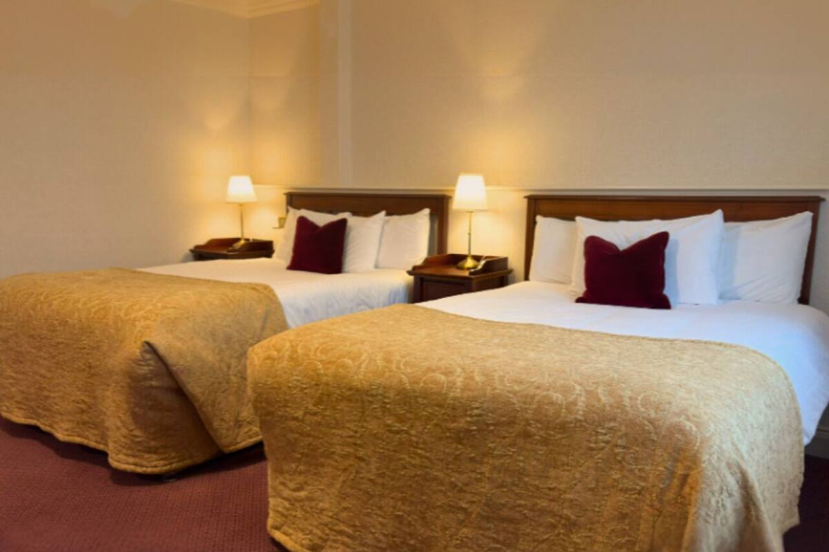 A hotel room with two beds, each with a wooden headboard, white linens, and a red pillow. There are bedside tables with lamps and a red carpet.