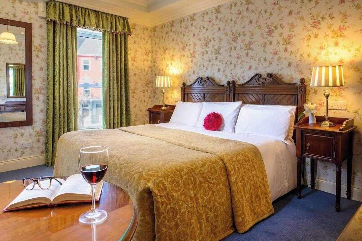 A hotel room with a bed, two bedside lamps, a window with curtains, a bedside table with a book and a glass of wine, and a mirror on the wall.