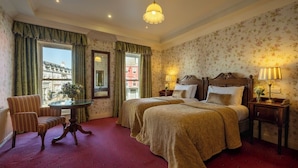 A hotel room with two beds, a sitting area with a chair and table, a lamp, and a view of buildings outside the window. - Arbutus Hotel Killarney (Killarney)