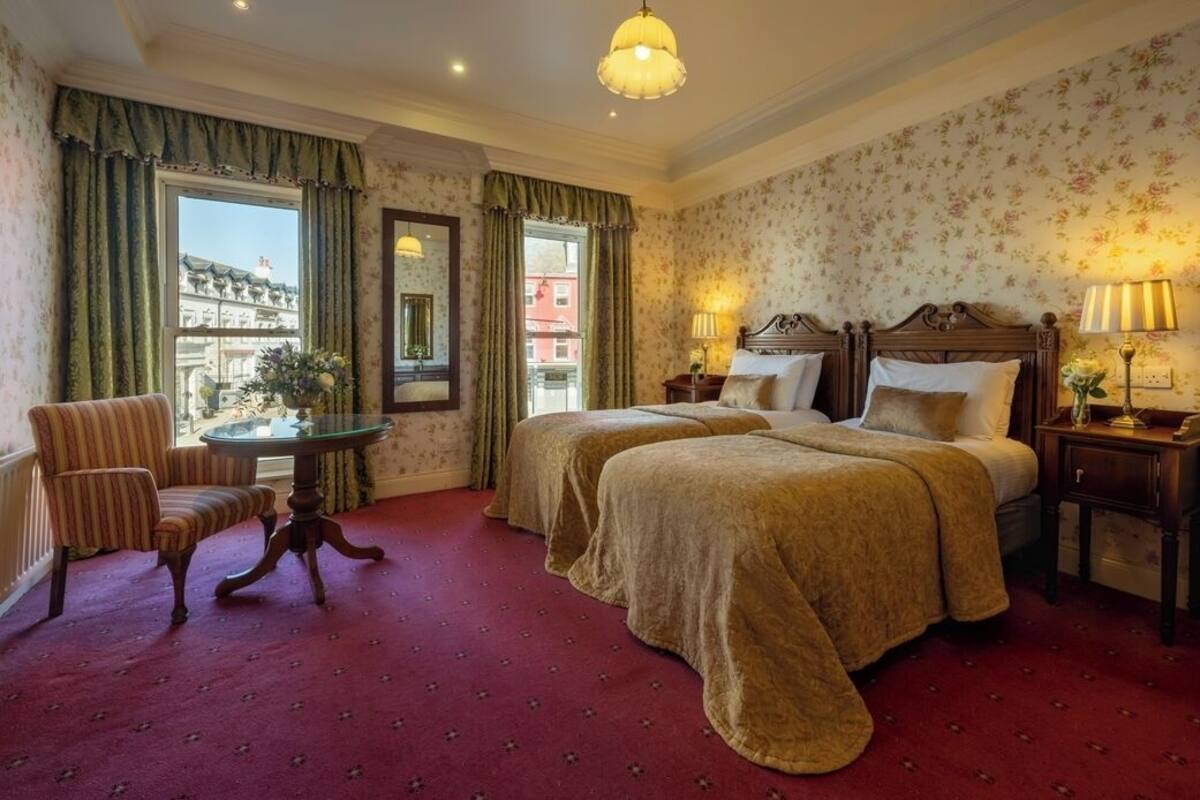 A hotel room with two beds, a sitting area with a chair and table, a lamp, and a view of buildings outside the window.