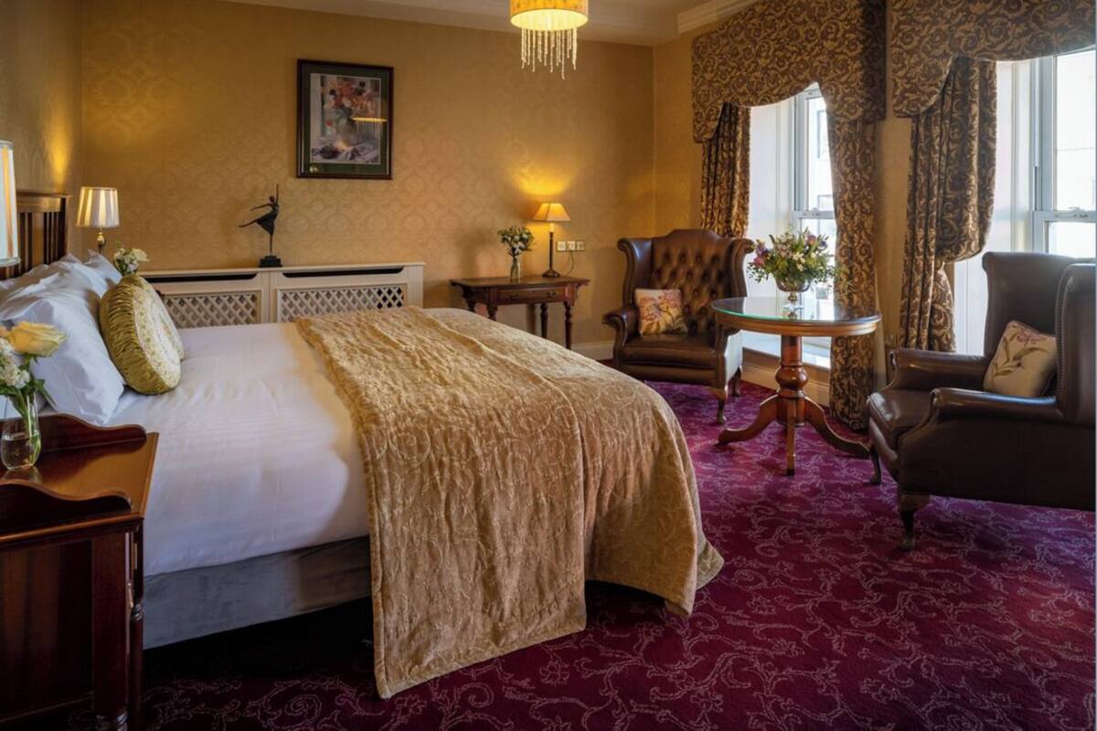 A hotel room with a large bed, a sitting area with a chair and table, a lamp, and a window with curtains.