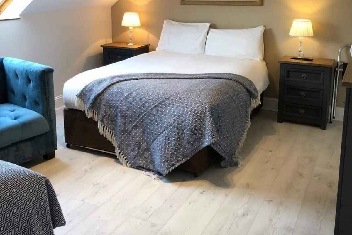 Quarto com cama, duas mesas de cabeceira, luminária, poltrona azul e um quadro na parede.