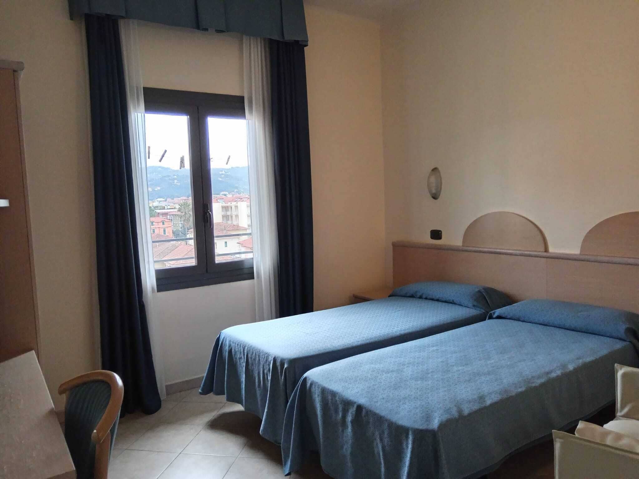 Une chambre d’hôtel avec deux lits simples, une fenêtre avec des rideaux, une chaise et une vue sur les bâtiments environnants.