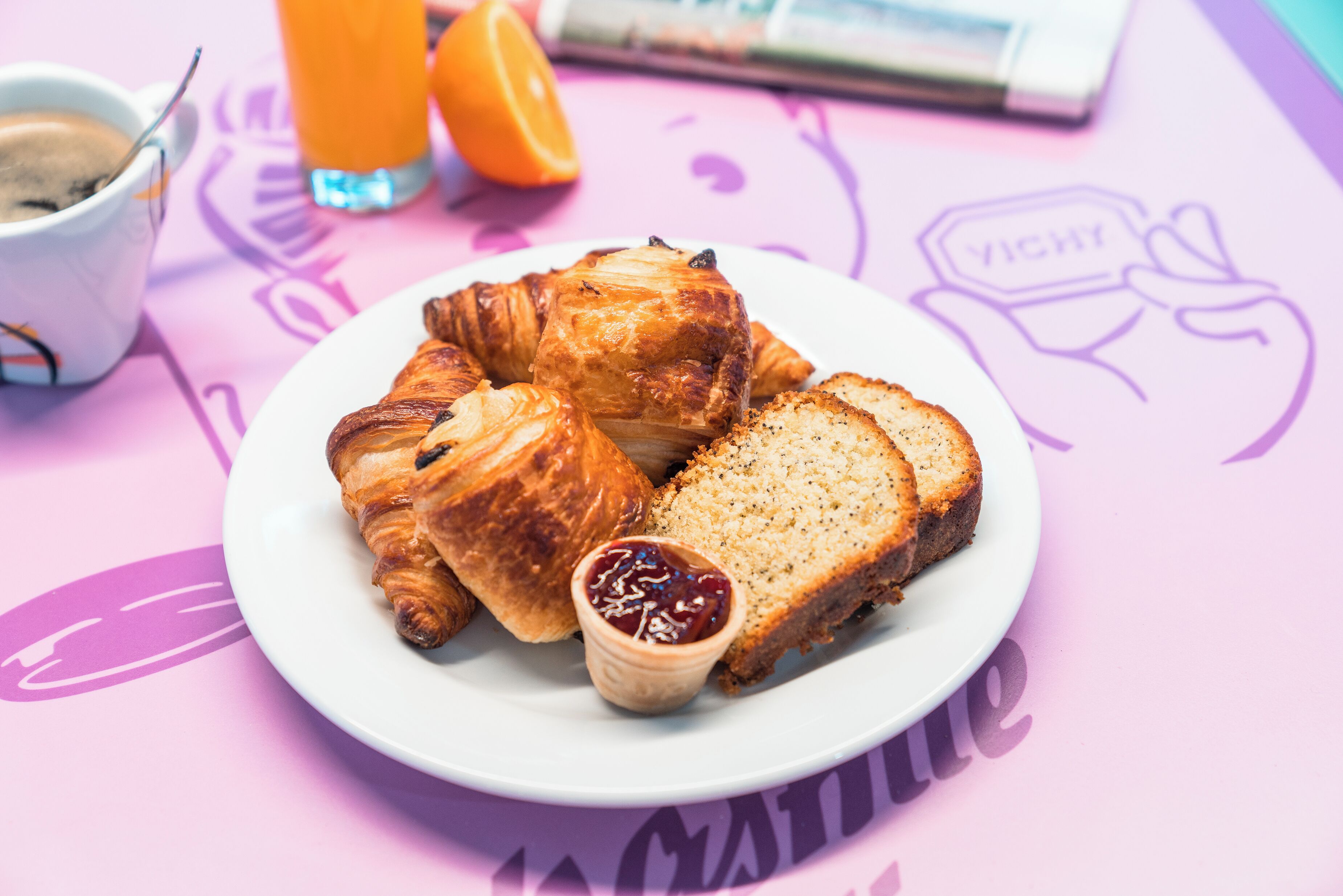 Een bord met croissants, een sneetje brood en een klein potje jam op een tafel met een tijdschrift en een kop koffie.