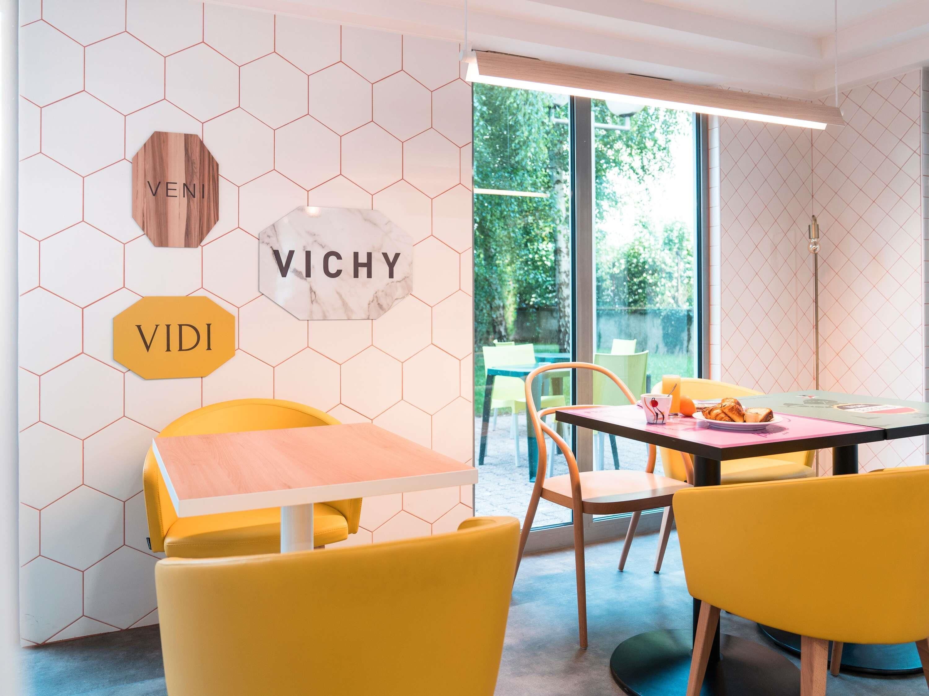 Un intérieur moderne avec des carreaux hexagonaux aux murs, un panneau en bois portant l’inscription « VENI », un panneau en marbre portant l’inscription « VICHY », et une chaise jaune.