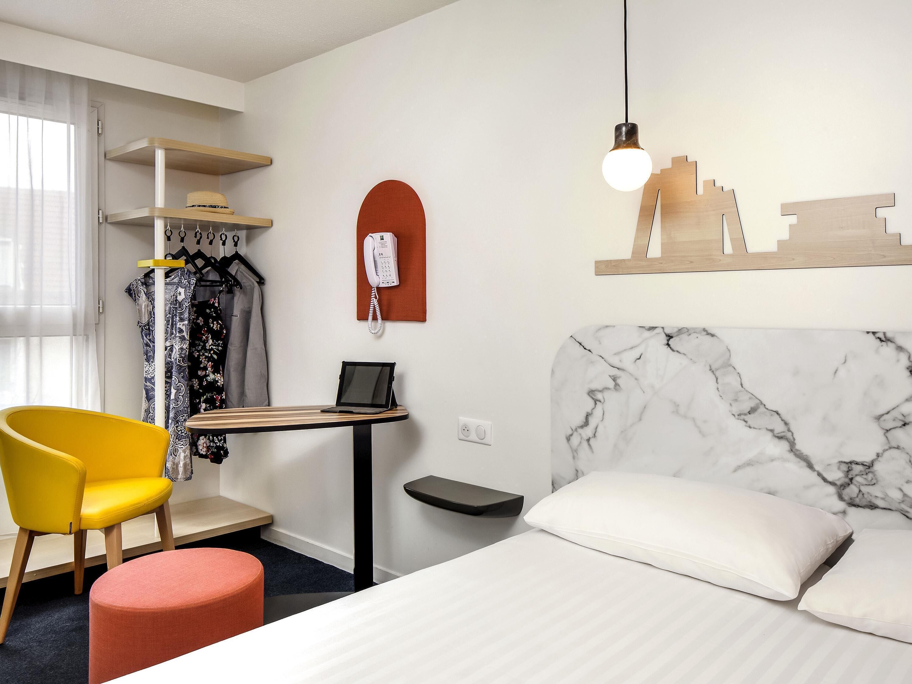 Une chambre d’hôtel comprenant un lit, un bureau avec une tablette, un téléphone, une chaise et un porte-manteau mural.