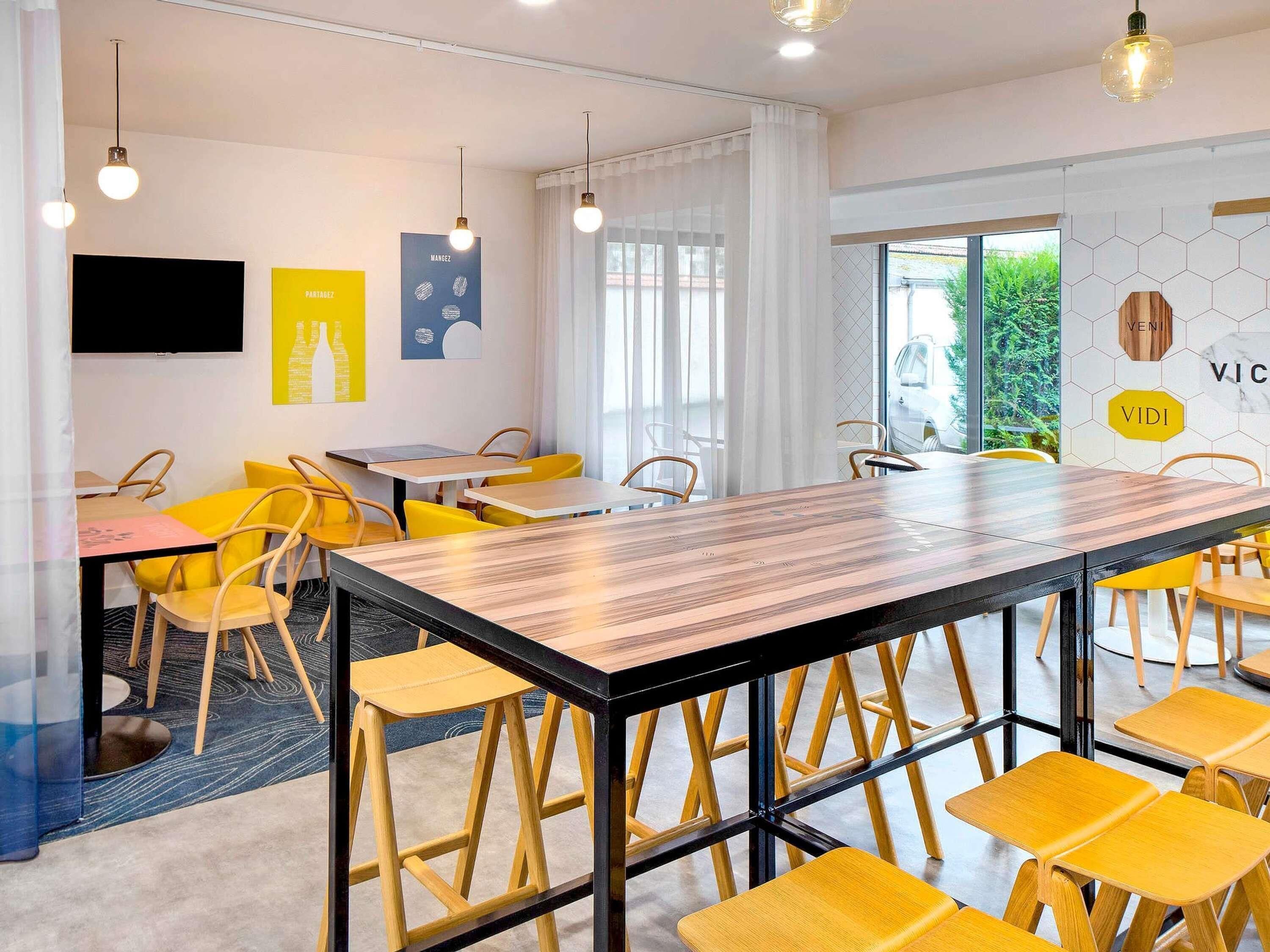 A modern dining area with a long wooden table, yellow chairs, and pendant lights.