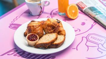 Un plato de desayuno con cruasanes, una taza de café y un vaso de zumo de naranja sobre una mesa rosa con una revista.