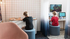 A modern room with two children using computers at a desk, a large clock on the wall, and a TV screen displaying an animated show. - ibis Styles Vichy Centre (Vichy)