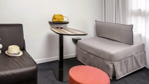 A modern hotel room with a grey sofa, a small round ottoman, a side table with a yellow bag and sunglasses, and a window with curtains. - ibis Styles Vichy Centre (Vichy)