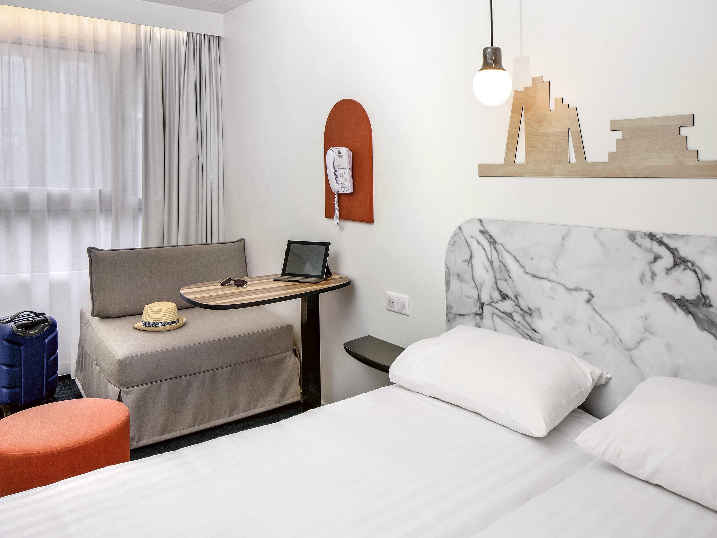 Ett hotellrum med en säng, ett skrivbord med en bärbar dator, en telefon och ett konstverk som föreställer en stadsbild på väggen.