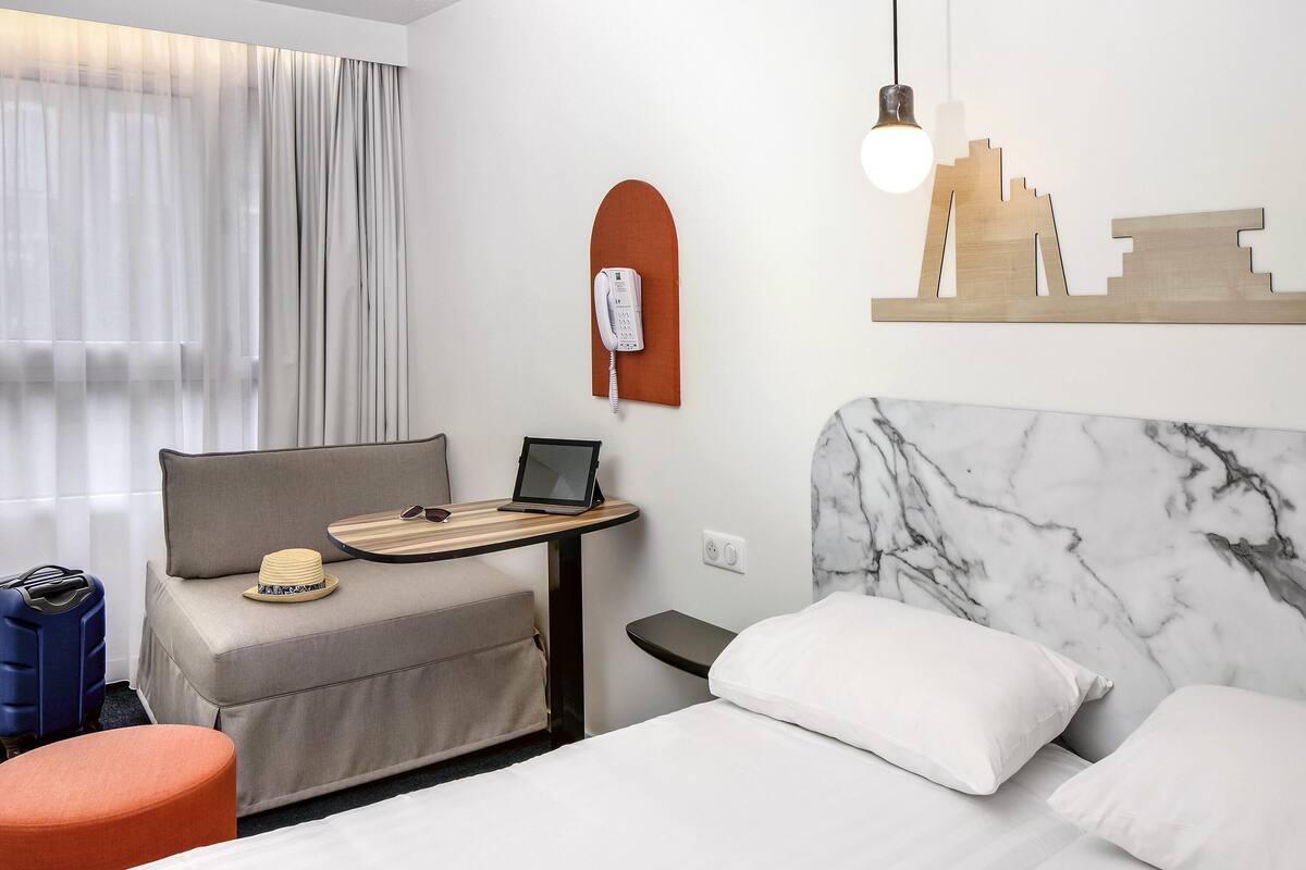 Ein Hotelzimmer mit einem Bett, einem Schreibtisch mit Laptop, einem Telefon und einem Stadtbild an der Wand.