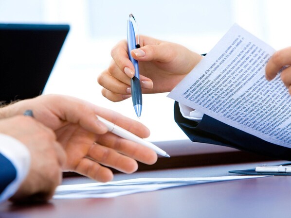 Una persona sosteniendo un bolígrafo y un documento, con otra mano extendiéndose, posiblemente en un entorno de negocios.