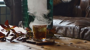 A glass of whiskey with smoke rising from it, placed on a wooden surface with a fur-covered chair in the background. - Hotel de la Poste - Relais Napoléon III (Bouillon)