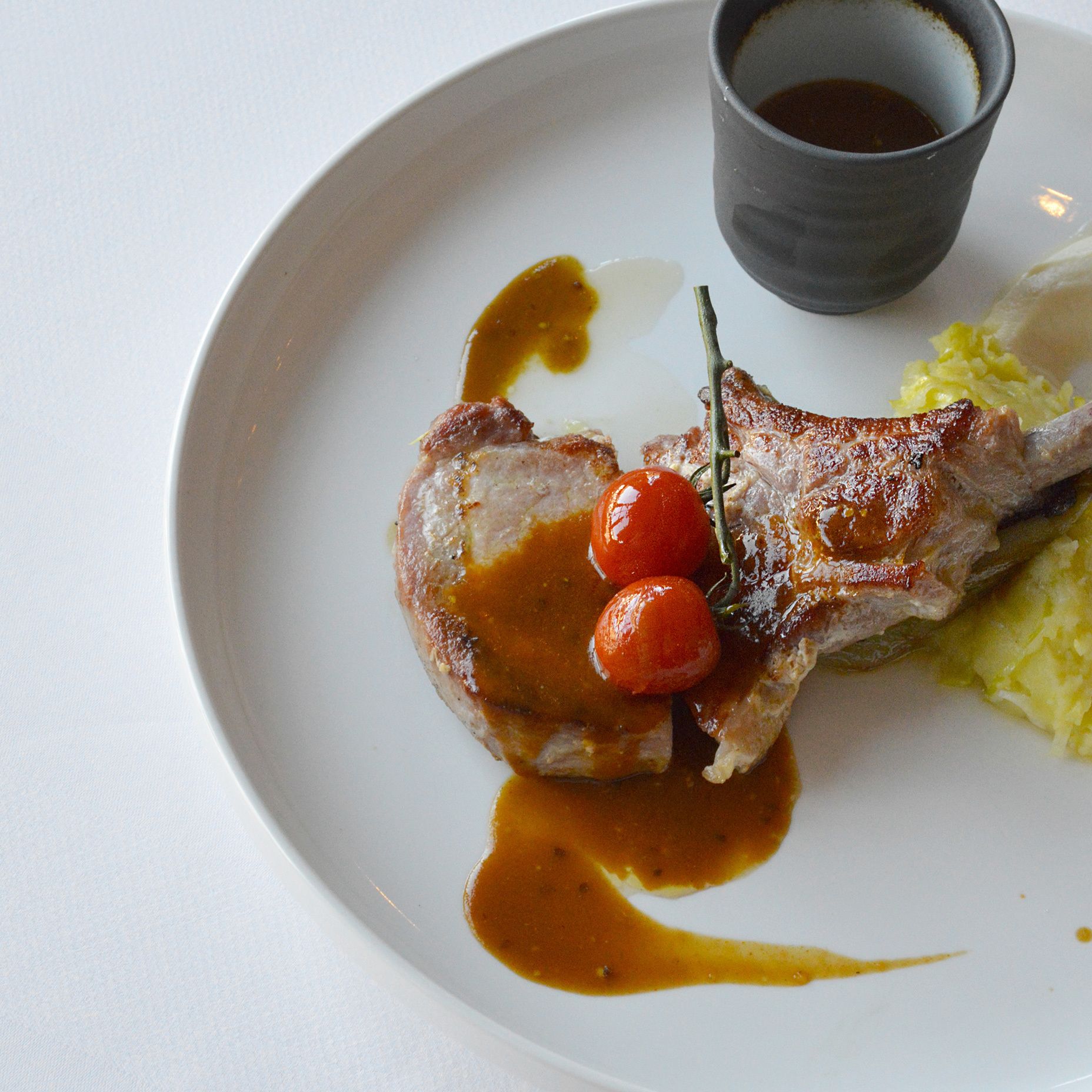 A plate with a lamb chop, cherry tomatoes, and a small cup of sauce.
