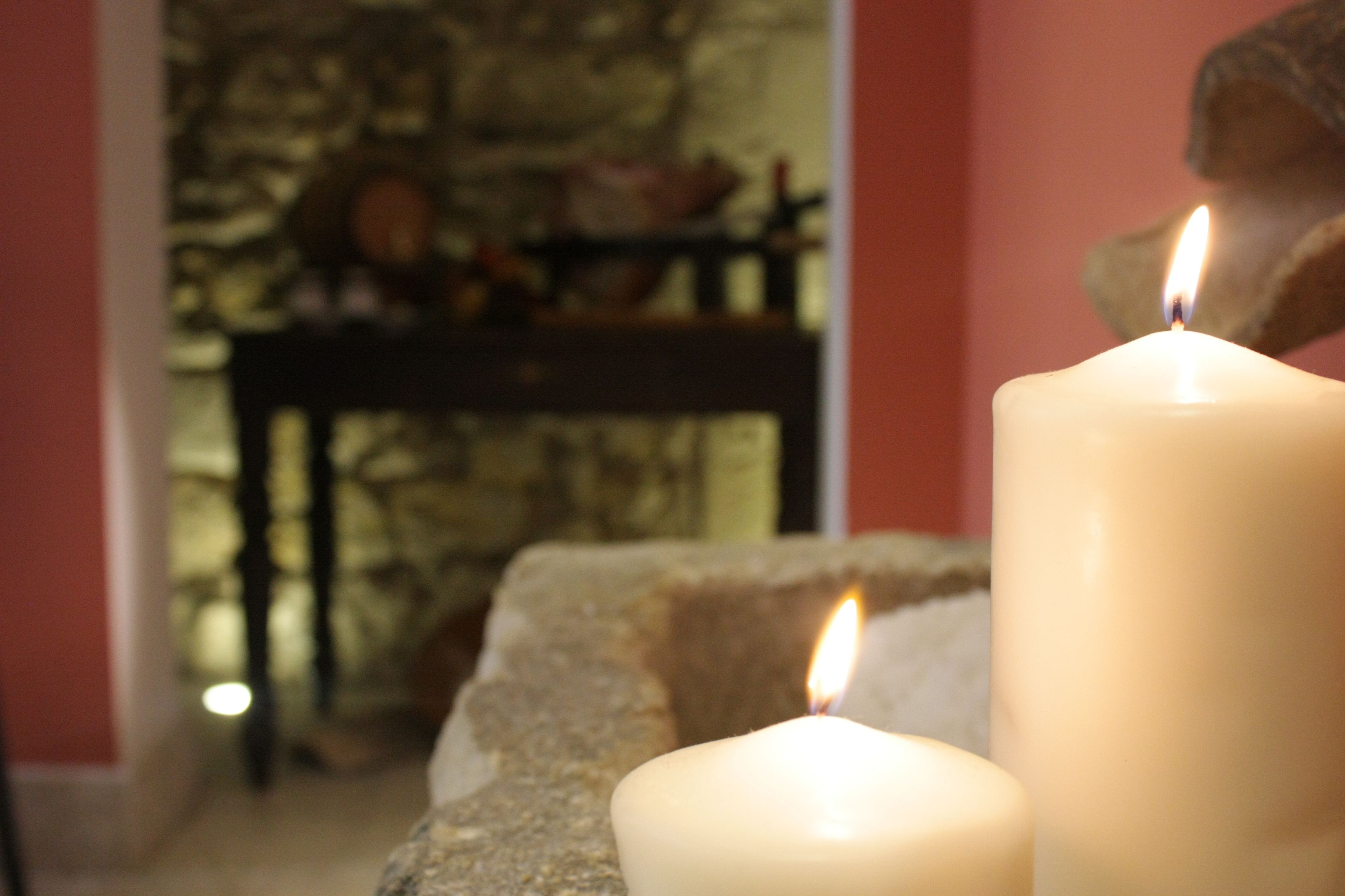 Two lit candles on a stone surface, with a blurred background including a stone wall and a wooden table with various items.