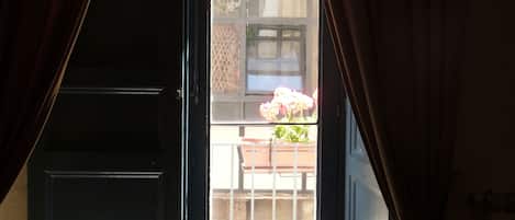 Una habitación con un ventanal, cortinas y vistas a un balcón con barandilla y plantas en macetas.
