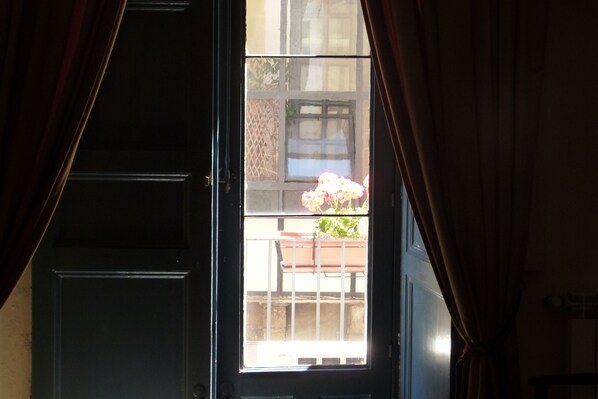 A room with a large window, curtains, and a view of a balcony with a railing and potted plants.