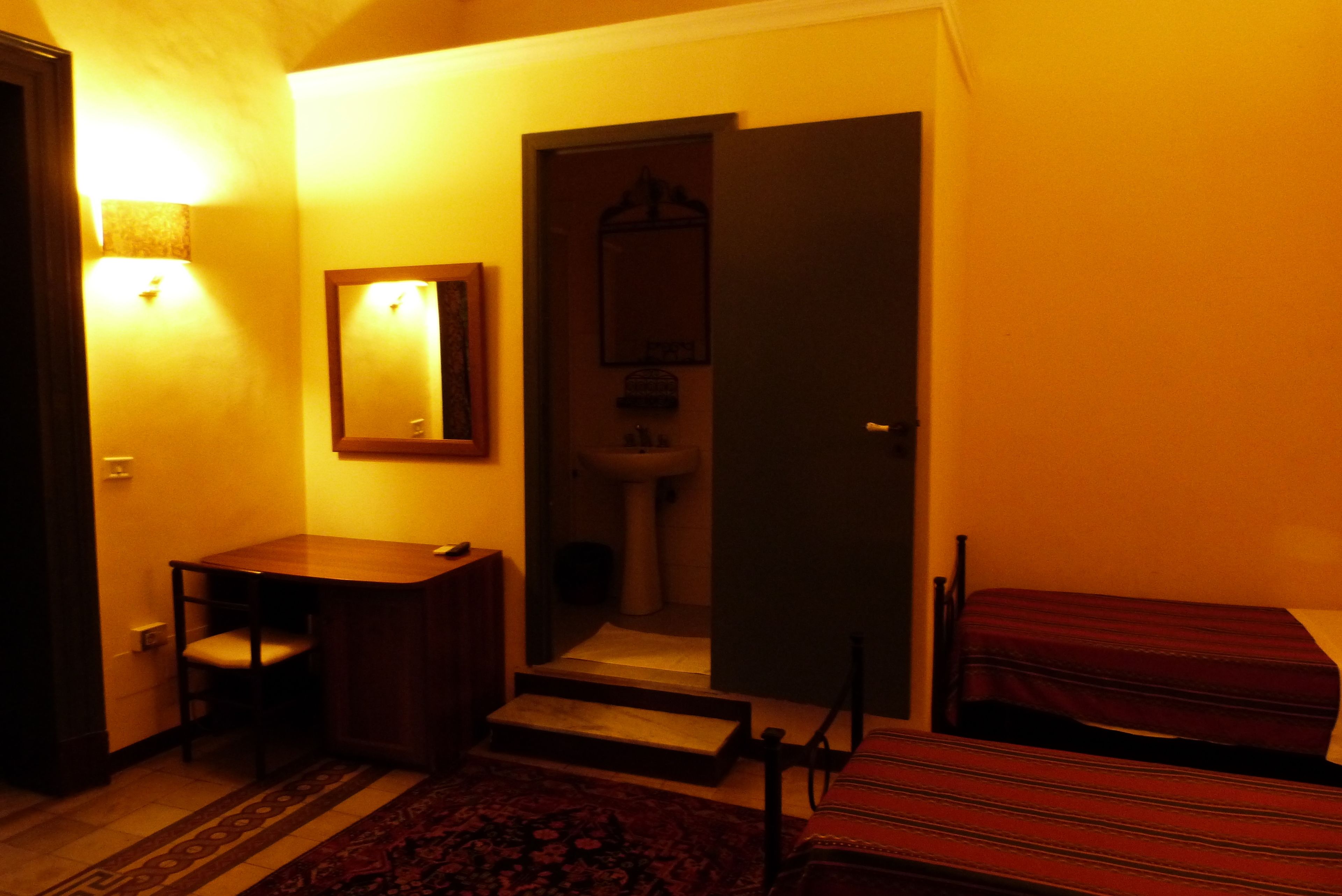 Um quarto pequeno e com pouca luz, com uma cama, uma cadeira, uma mesinha e um espelho na parede.