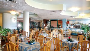A spacious restaurant with high ceilings, wicker chairs, and tables set with blue tablecloths. - Sporting Baia Hotel (Giardini Naxos)