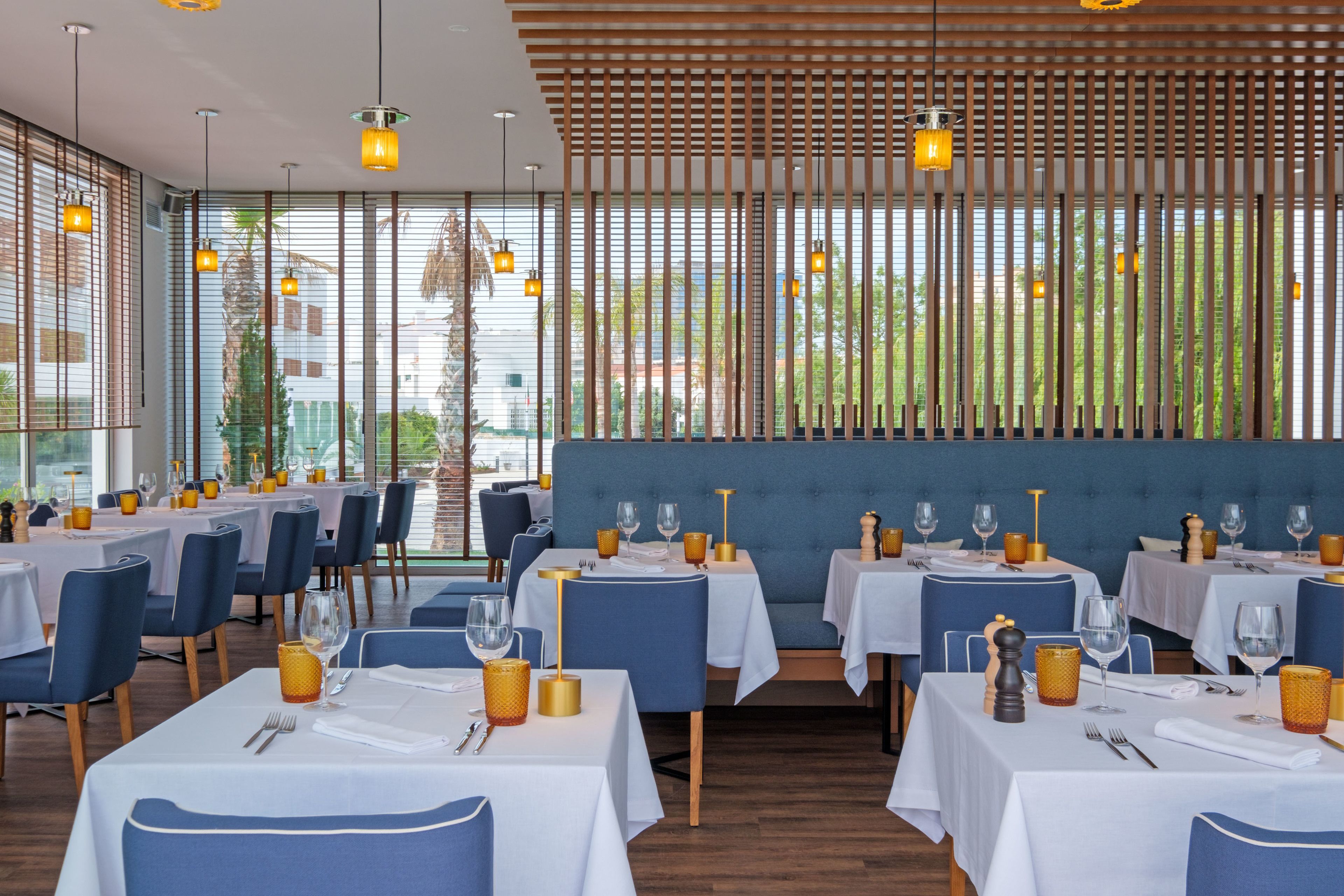 Um restaurante com várias mesas, assentos estofados azuis e vista para o verde lá fora.