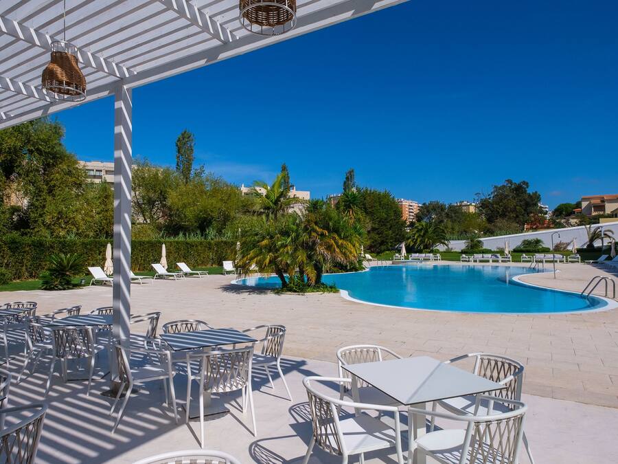 Un área junto a la piscina con mesas y sillas blancas, cielo azul despejado y una piscina al fondo.