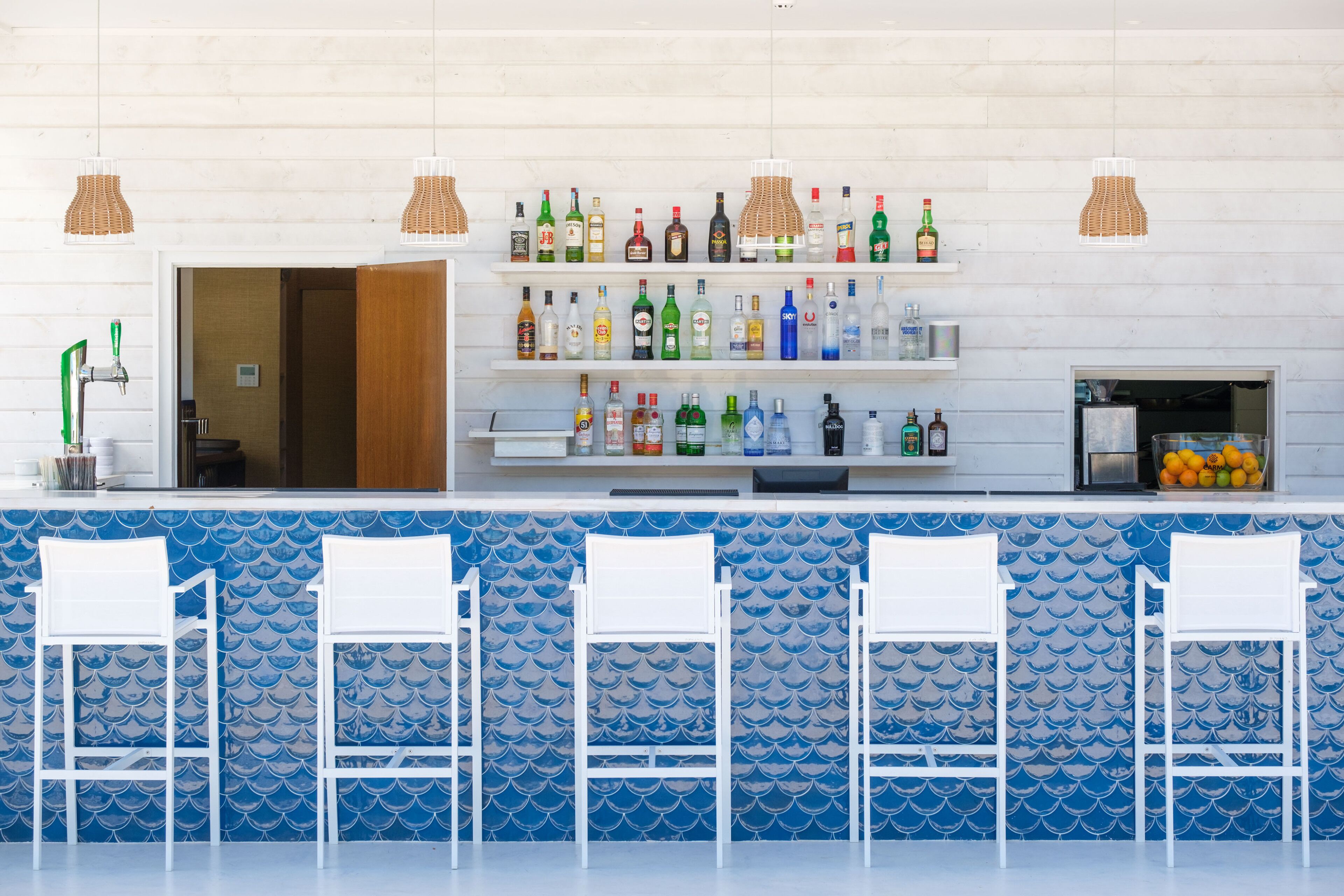 A bar area with a blue tiled backsplash, white bar stools, and shelves stocked with various bottles.