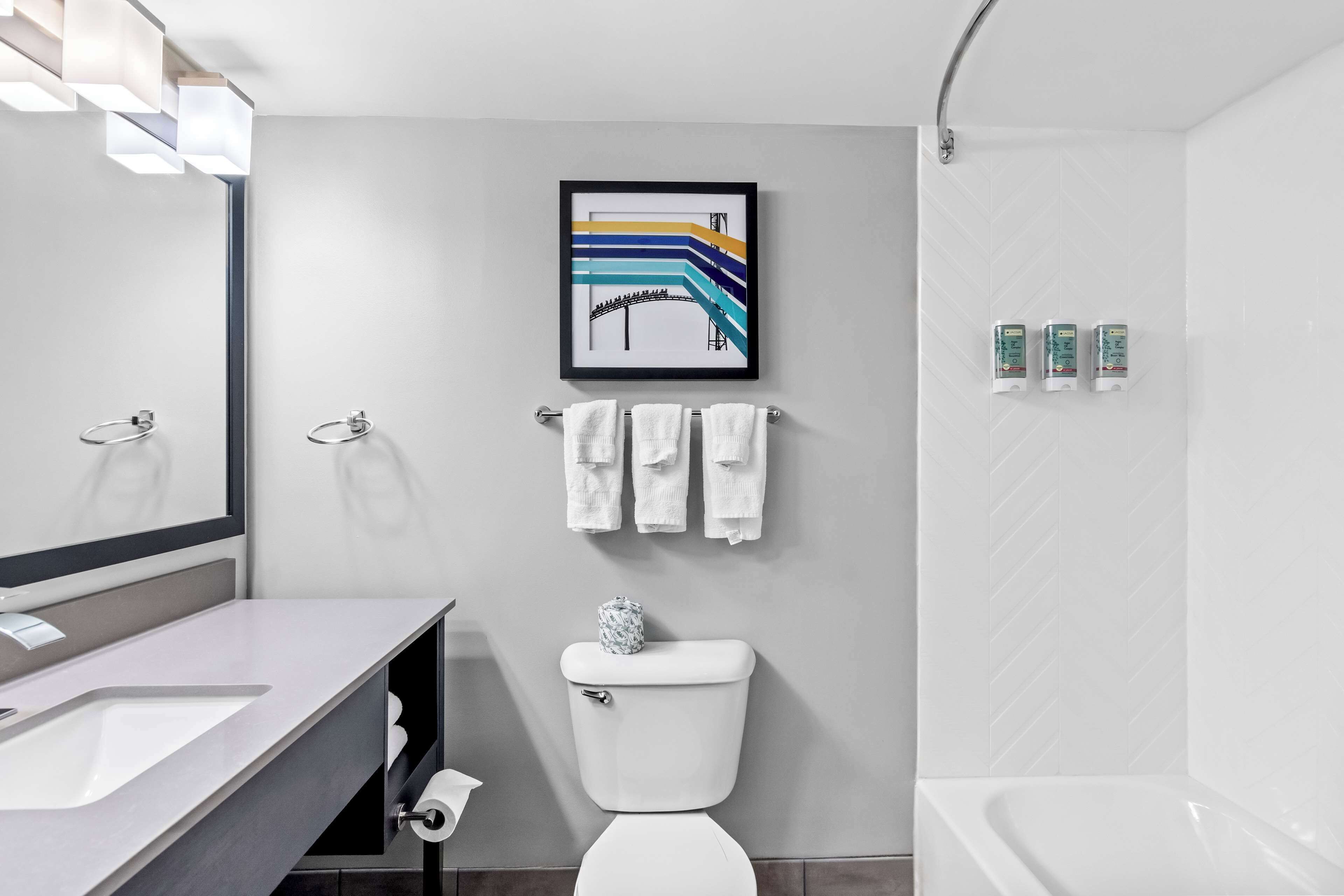 Une salle de bain moderne avec un lavabo, des toilettes et une baignoire, tous de couleur blanche. Un tableau encadré est accroché au-dessus des toilettes, et un porte-serviettes contient des serviettes.