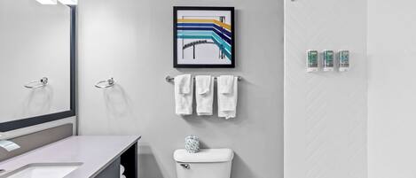 Une salle de bain moderne avec un lavabo, des toilettes et une baignoire, tous de couleur blanche. Un tableau encadré est accroché au-dessus des toilettes, et un porte-serviettes contient des serviettes.