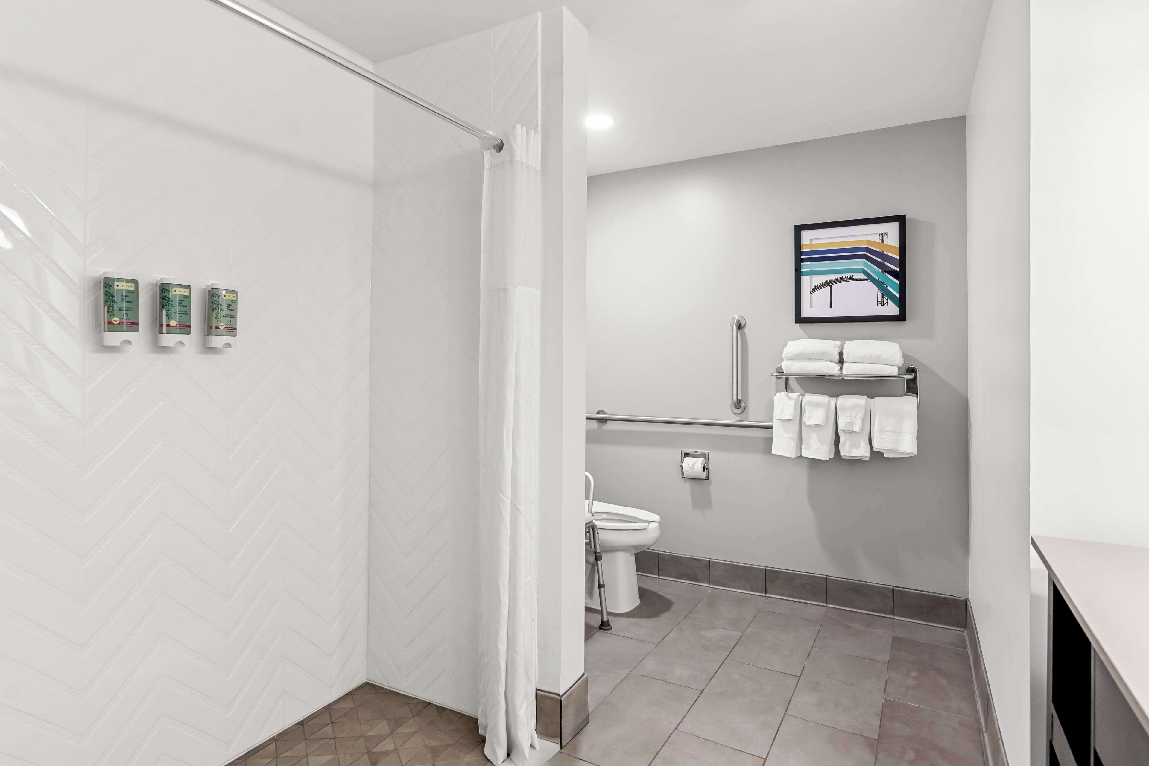 Un baño moderno con ducha, inodoro y barras de apoyo. Hay tres secamanos fijos en la pared.
