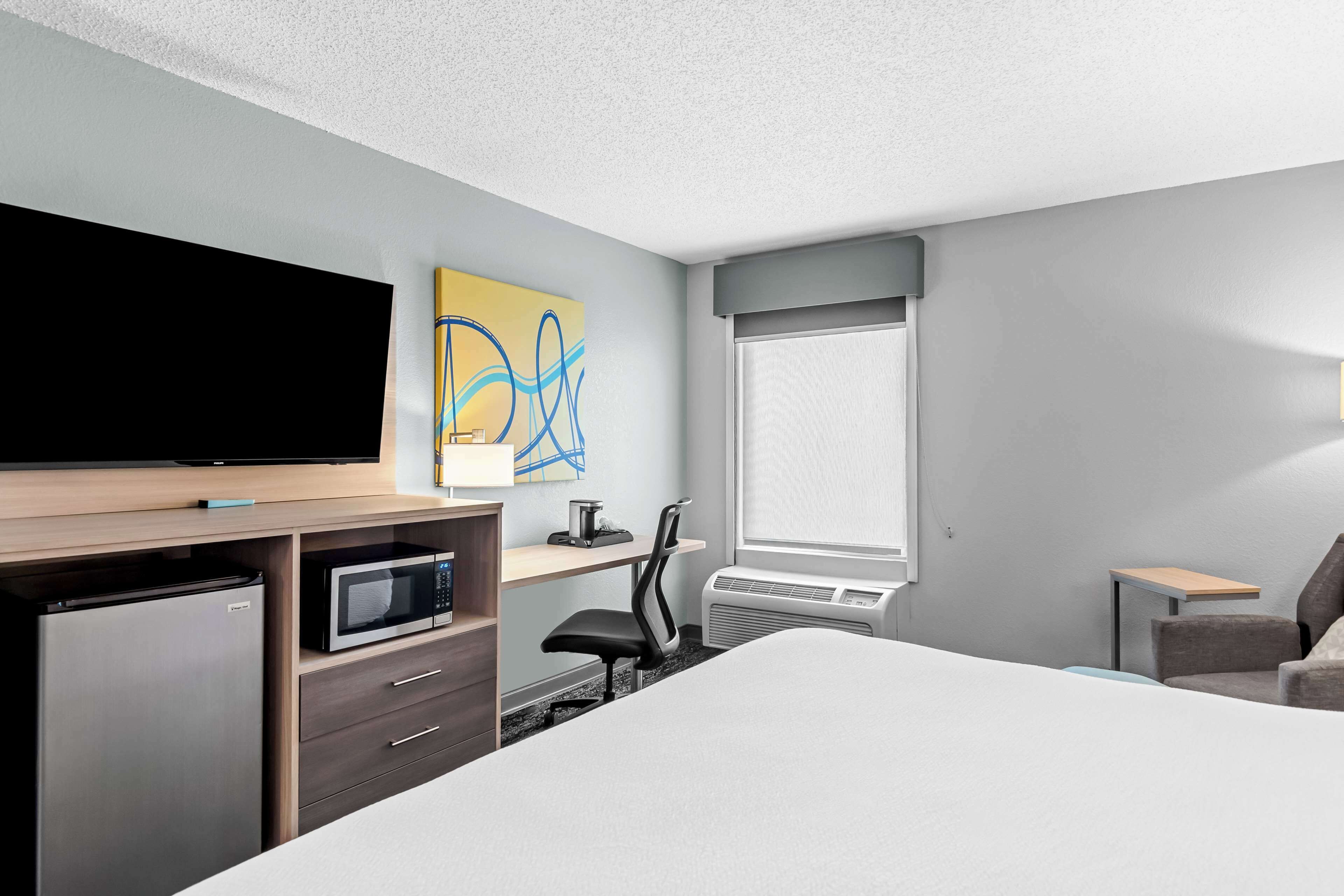 Habitación de hotel con cama, escritorio con computadora, microondas, refrigerador, televisor y ventana con persianas.