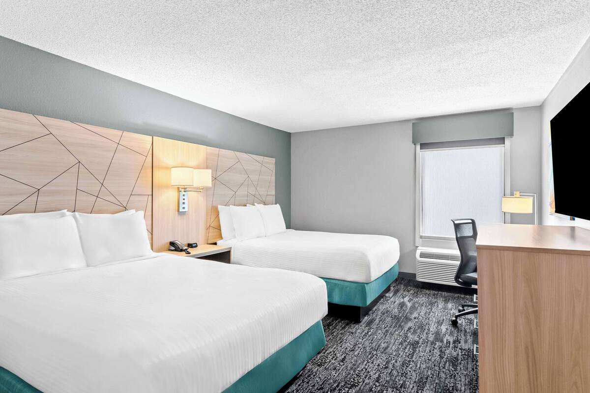 A hotel room with two beds, a desk, a chair, a lamp, and a window.