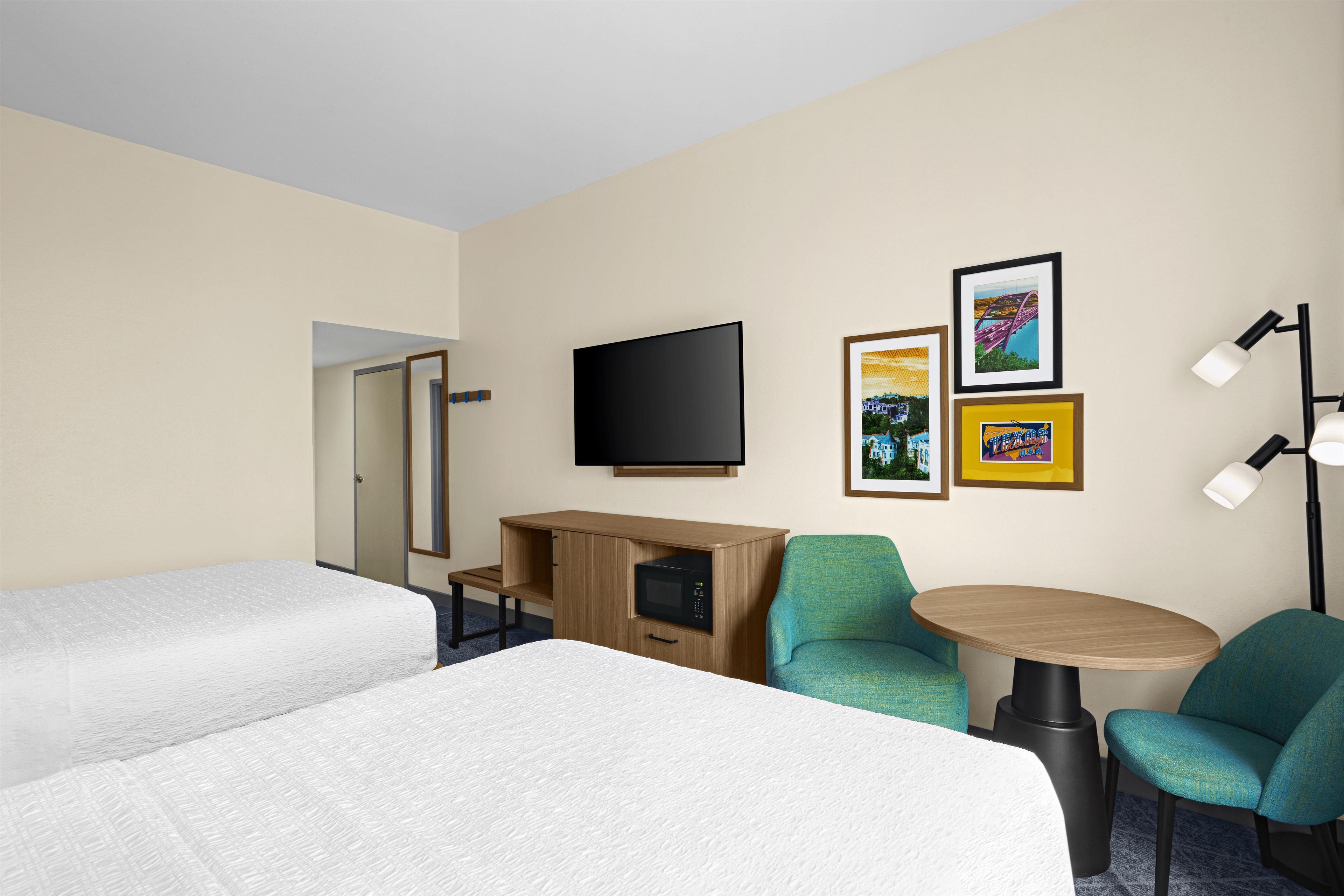 A hotel room with two beds, a desk with a TV, a small table, two chairs, and wall art.