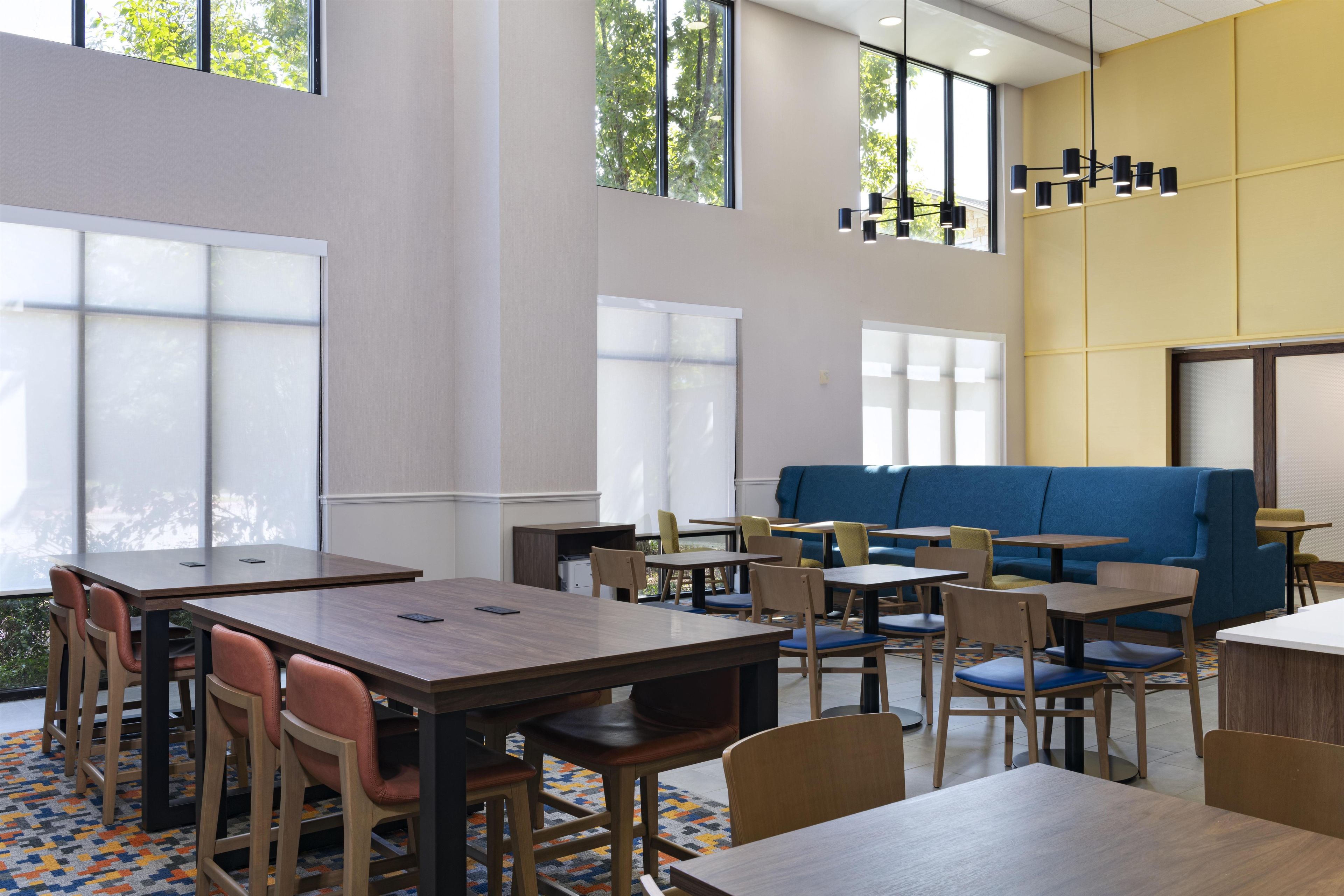 A modern dining area with tables and chairs, a blue sofa, and large windows allowing natural light.