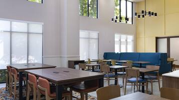 A modern dining area with tables and chairs, a blue sofa, and large windows allowing natural light.