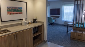 Une chambre d’hôtel moderne équipée d’une kitchenette, d’un téléviseur à écran plat, d’une grande fenêtre avec des rideaux et d’un tableau encadré au mur.
