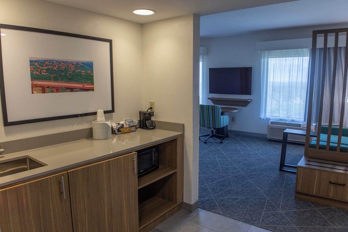 A modern hotel room with a kitchenette, a flat-screen TV, a large window with curtains, and a framed picture on the wall.