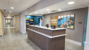 Un comptoir d’accueil avec une pancarte indiquant « Bienvenue à Hampton », une affiche présentant des photos des lieux et un couloir menant aux chambres.