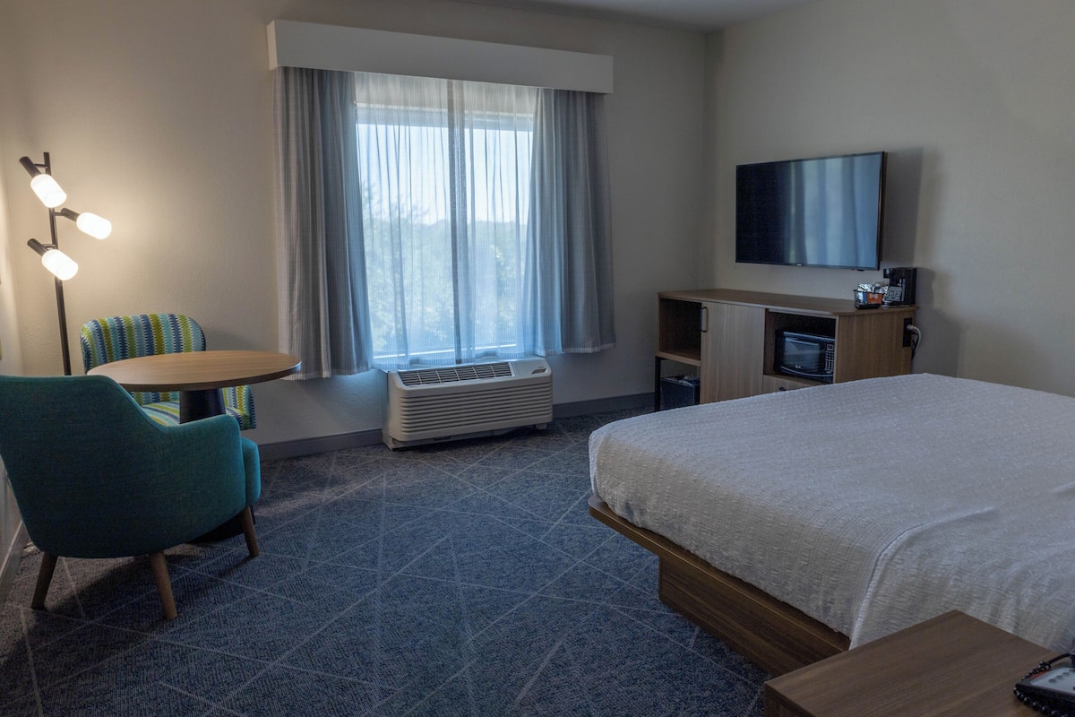 A hotel room with a bed, a desk with a chair, a TV, an air conditioner, and a window with curtains.