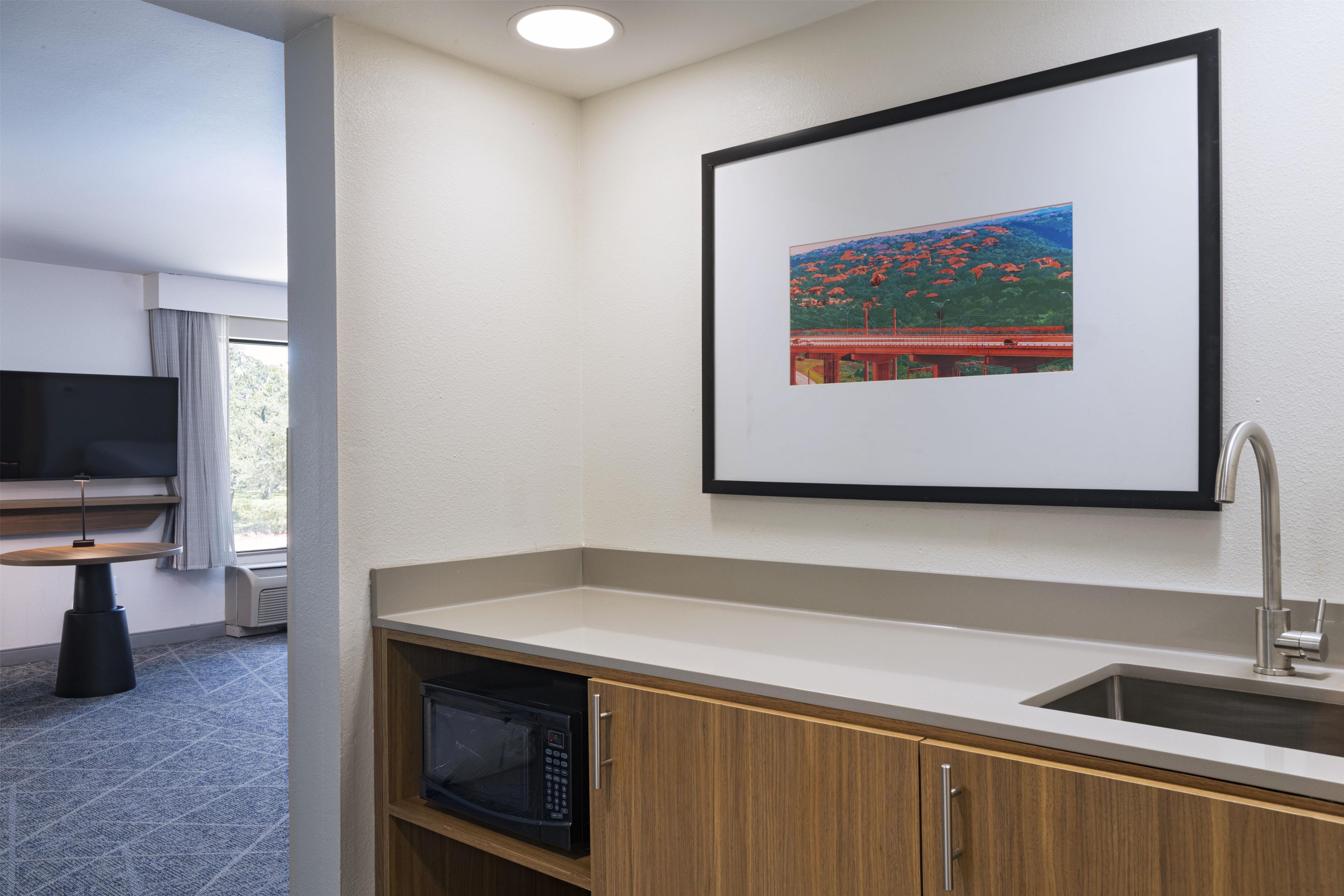 Ein Hotelzimmer mit Küchenzeile, einem Flachbildfernseher, einem Fenster mit Vorhängen und einem gerahmten Bild an der Wand.
