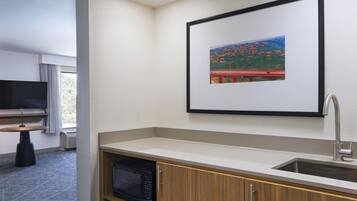 A hotel room with a kitchenette, a flat-screen TV, a window with curtains, and a framed picture on the wall.