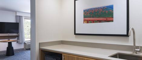 A hotel room with a kitchenette, a flat-screen TV, a window with curtains, and a framed picture on the wall.