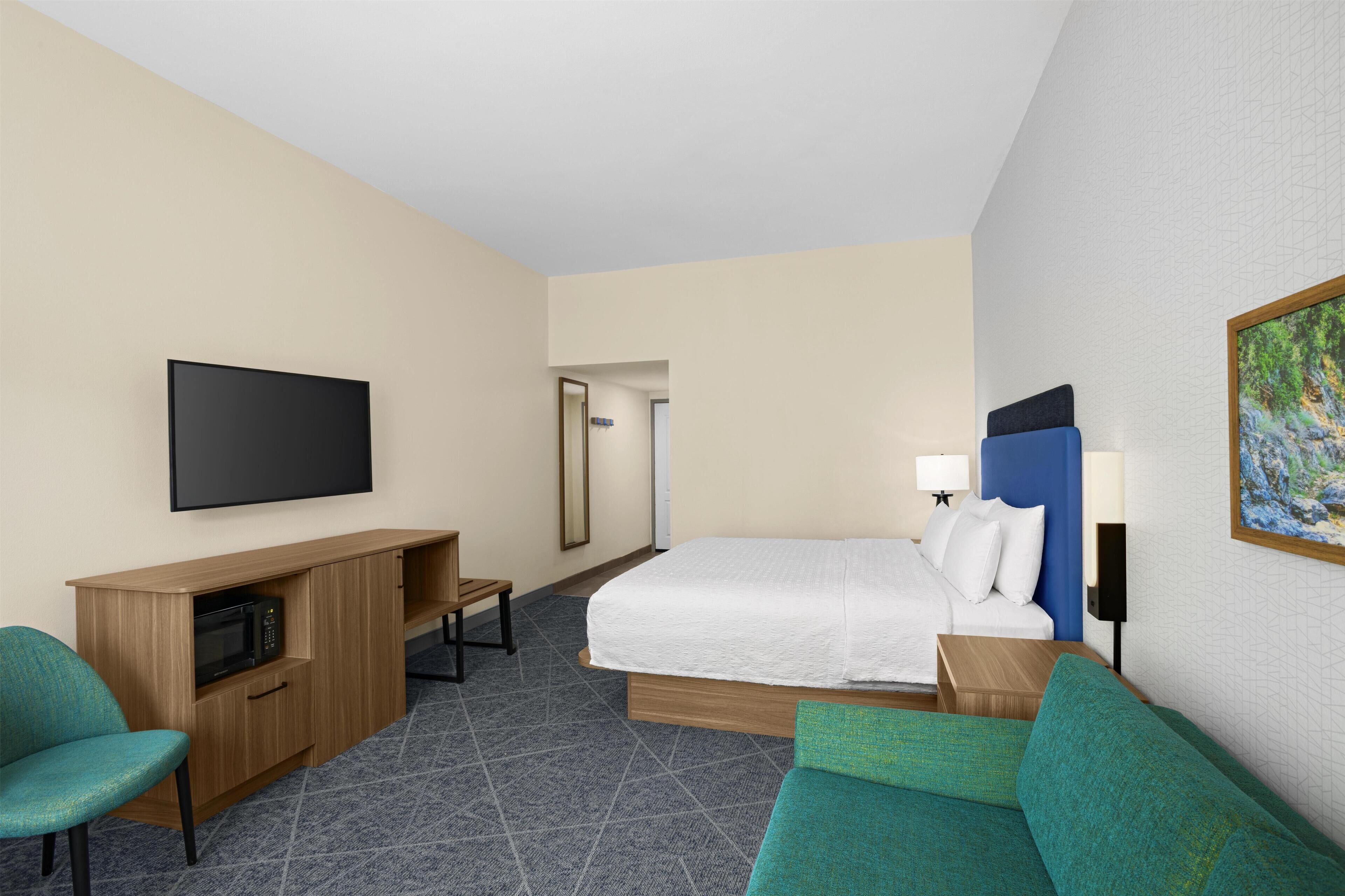 A hotel room with a bed, a TV, a desk, a chair, and a painting on the wall.